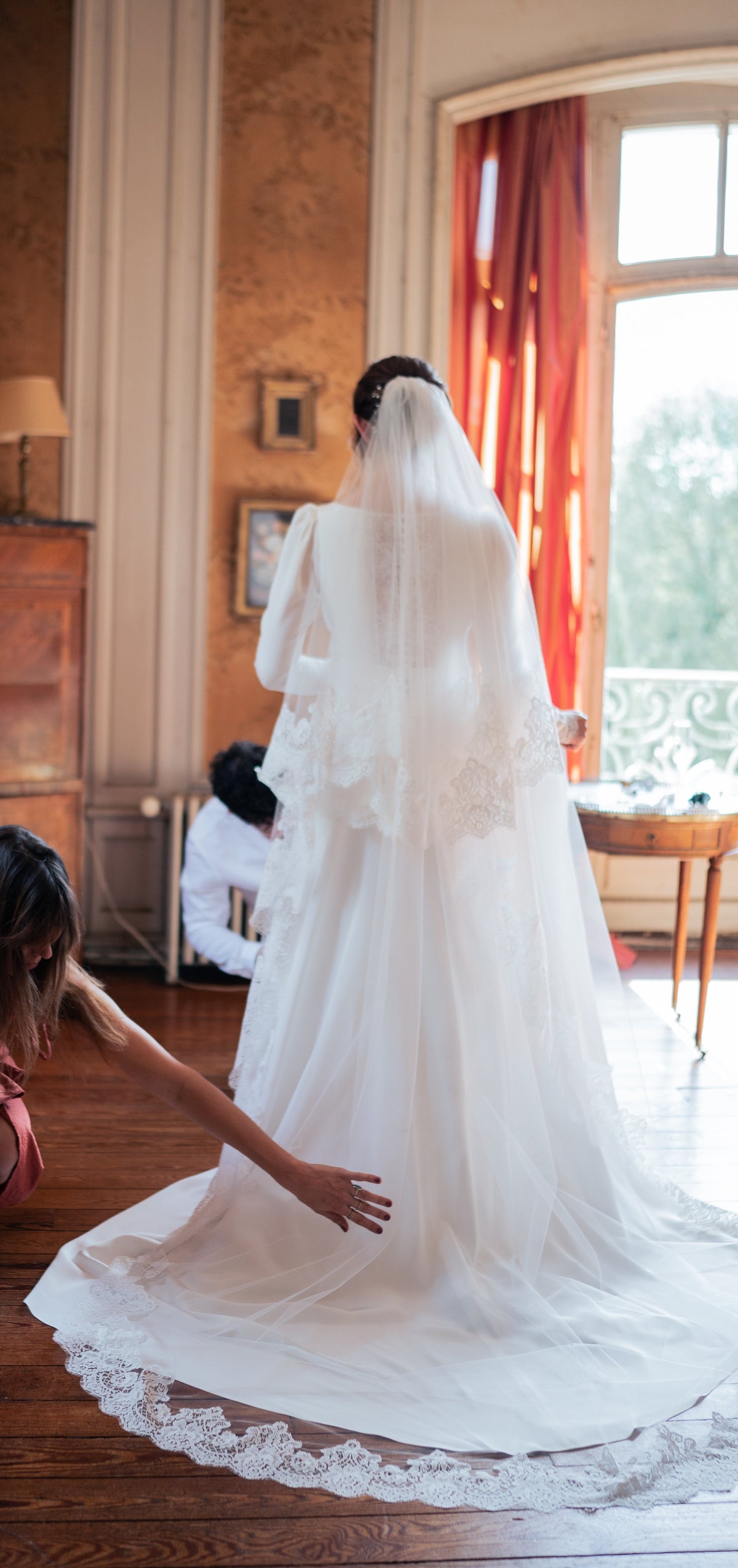 Bride seen from behind in a château room as a helper arranges her long lace-edged wedding veil