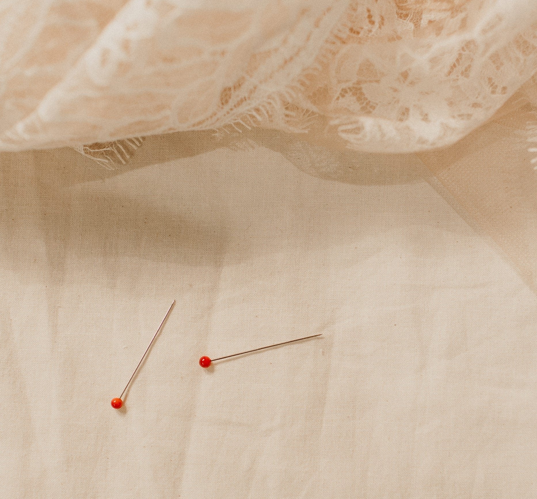 Delicate lace fabric placed on a beige surface with two red-headed sewing pins, evoking a handcrafted or bridal design process.
