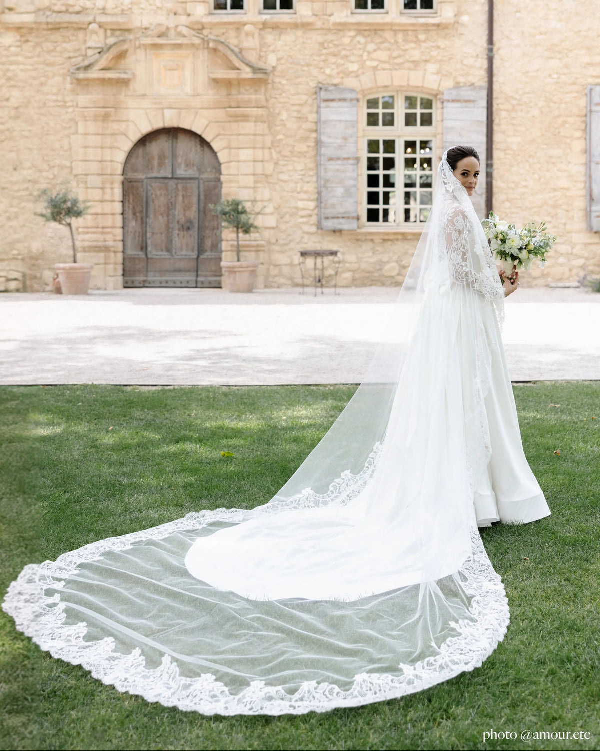 Bride on a lawn before a stone house, the Avec Toi lace mantilla spread across the grass