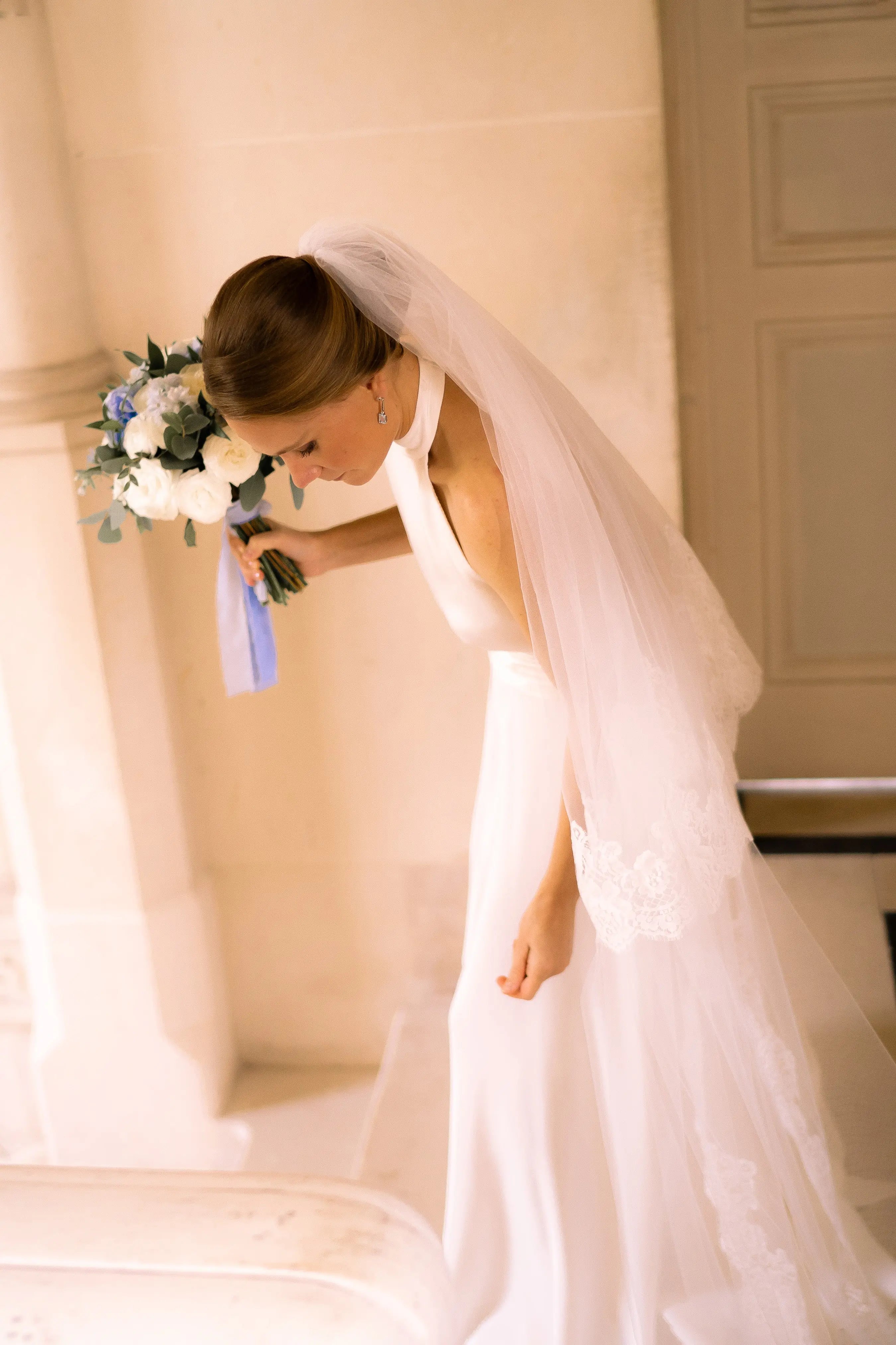 Bride looking at the ground as she holds her white, green and blue bouquet. She is wearing a halterneck wedding dress and a long wedding veil in her hair.