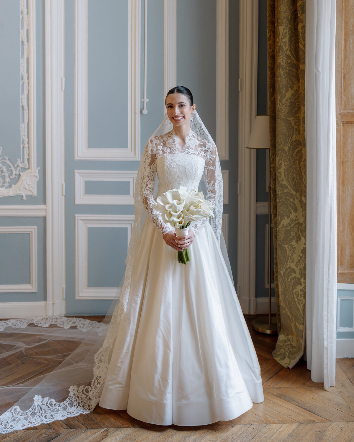 Bride holding calla lilies in a panelled room, wearing the Avec Toi long lace mantilla