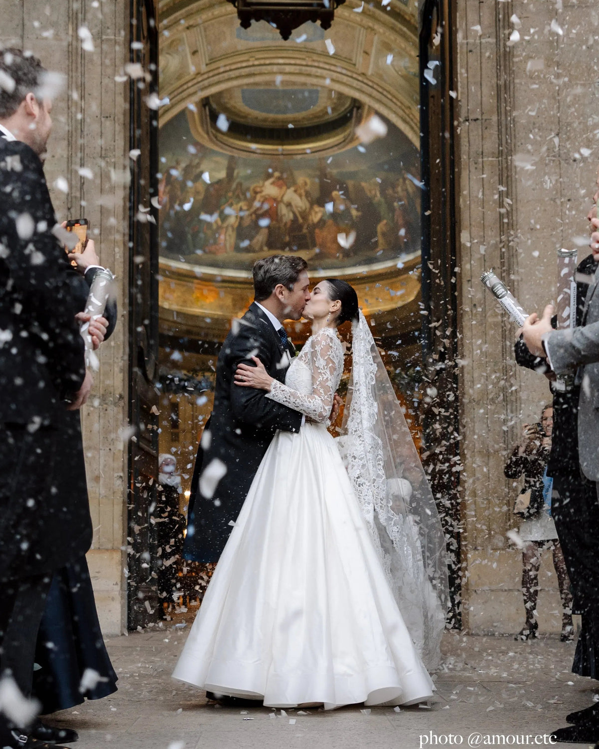 Bride and groom kissing at the church door under falling petals, the bride in the Avec Toi lace mantilla