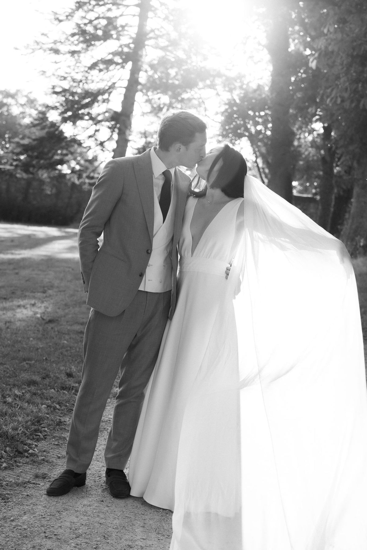 Black and white photo of a bride and groom kissing outdoors, her long silk chiffon veil blowing in the wind