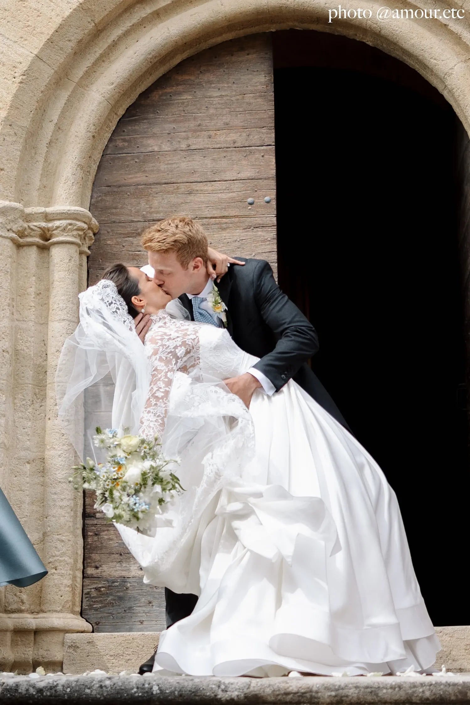 Groom kissing bride, wearing an ivory cathedral bridal mantilla veil in fine tulle with elegant lace edge.