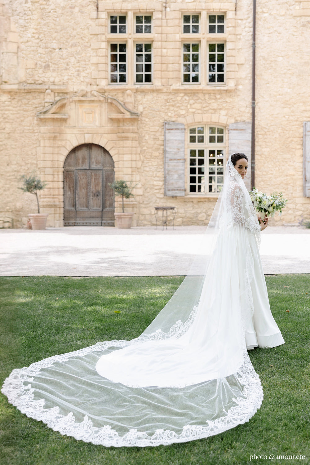 Bride turning her back to the camera and looking over her shoulder, her cathedral veil with a wide lace edge running from her face to a train spread across the lawn in front of a stone château