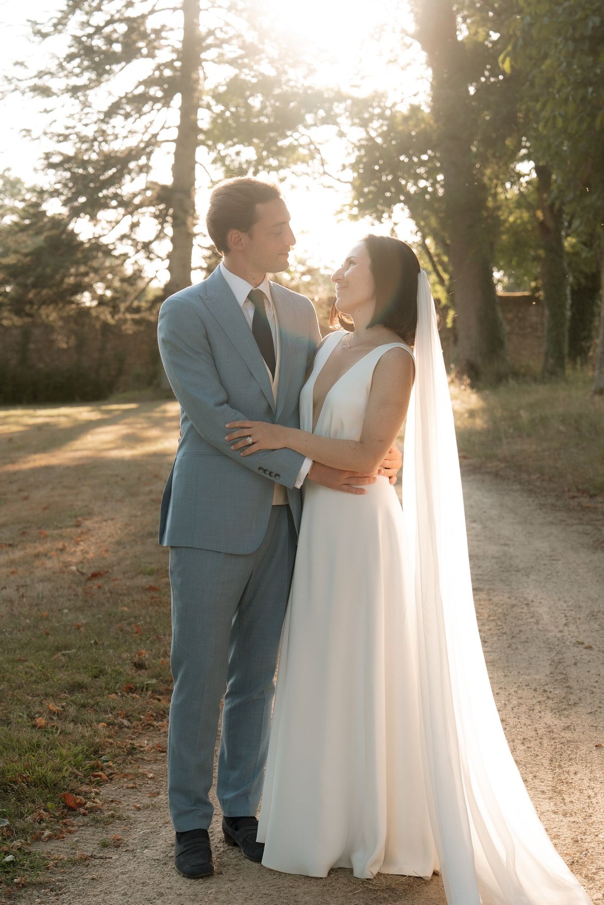 Bride and groom embracing in a sunlit park, her chapel-length silk chiffon veil falling to the ground behind her V-neck gown