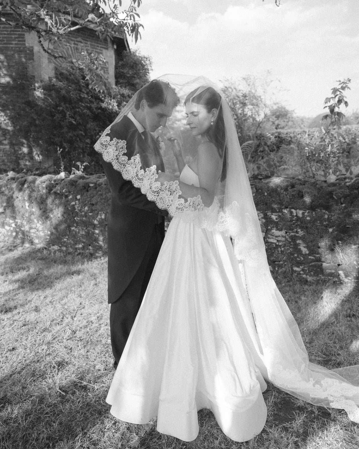 Black and white photo of a bride and groom standing under a long lace-edged wedding veil draped over both their heads in a garden