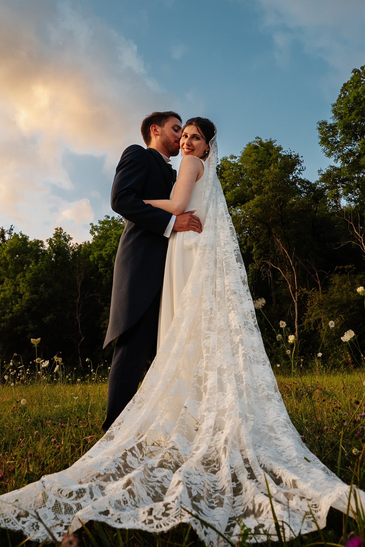 Bride and groom embracing in a meadow at sunset, her lace veil spreading across the grass
