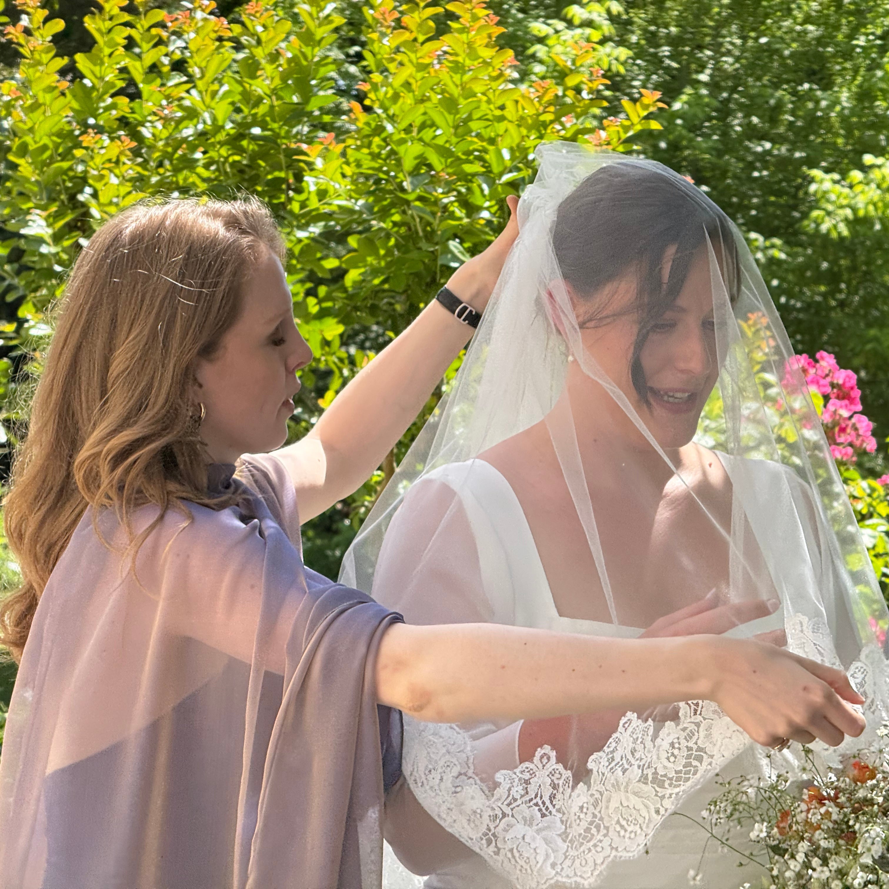 Lucy from The French Wedding Veil adjusting a bride's lace-trimmed wedding veil outdoors before the ceremony