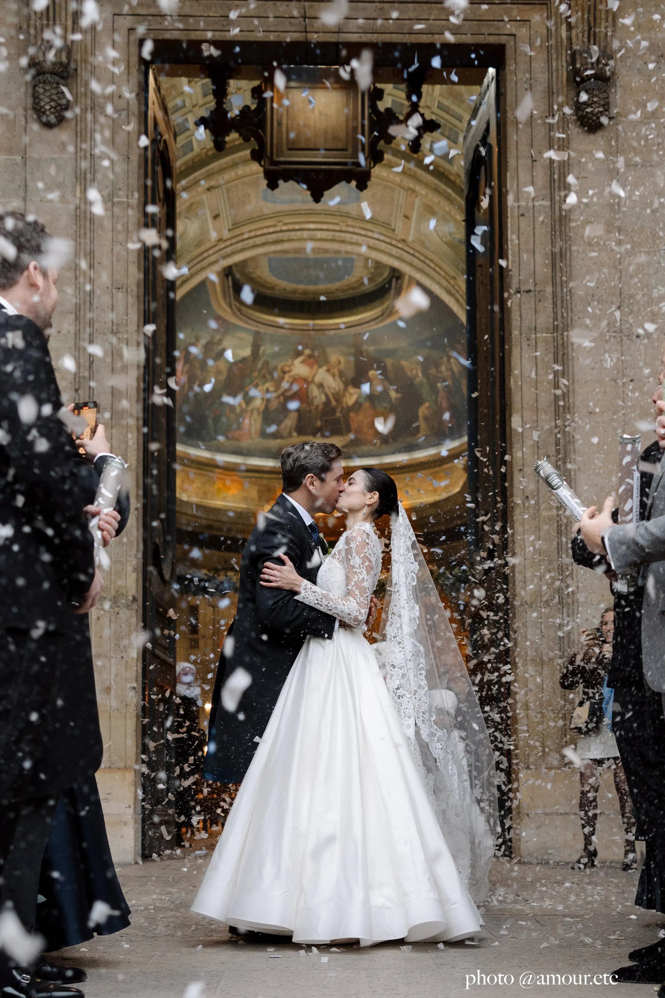 Bride and groom kissing in front of a church, the bride is wearing a cathedral bridal veil in fine tulle with elegant lace edge worn as a mantilla.