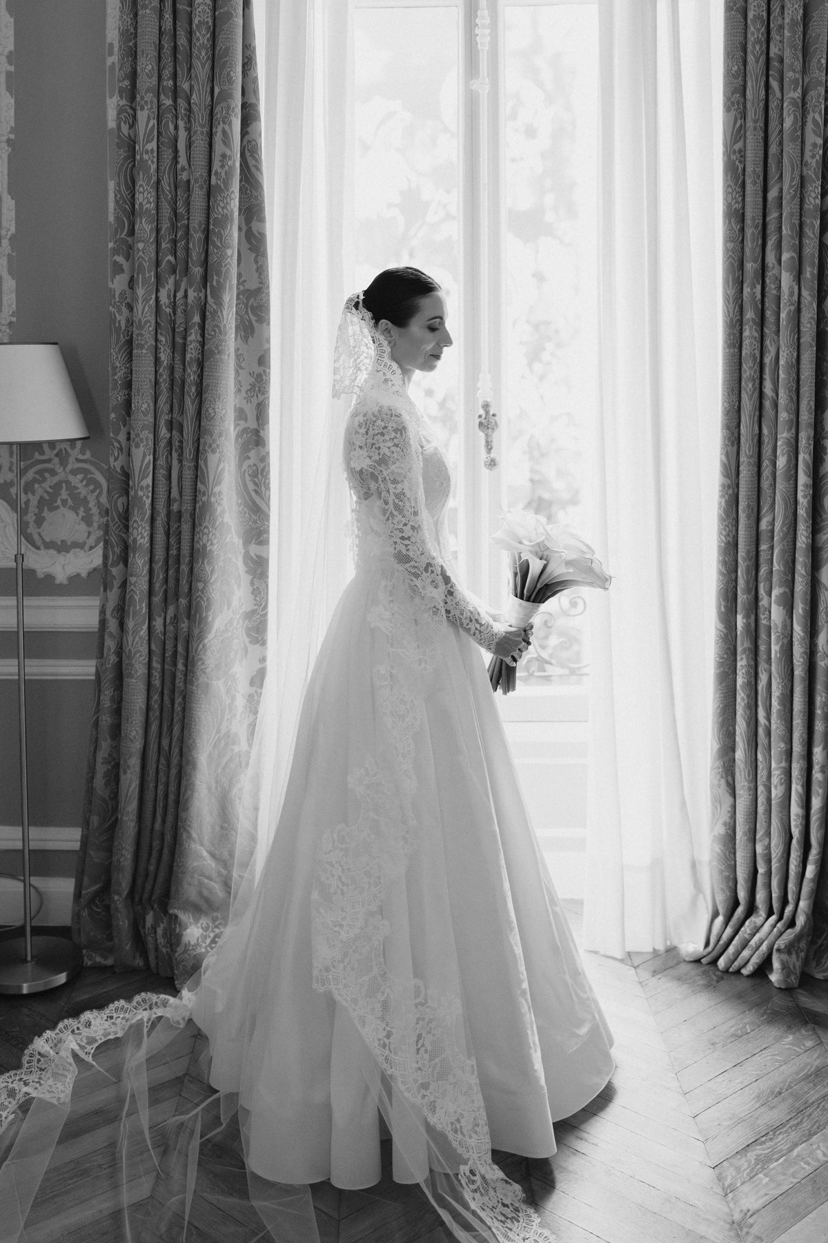Bride by a tall curtained window in the Avec Toi lace mantilla, in black and white