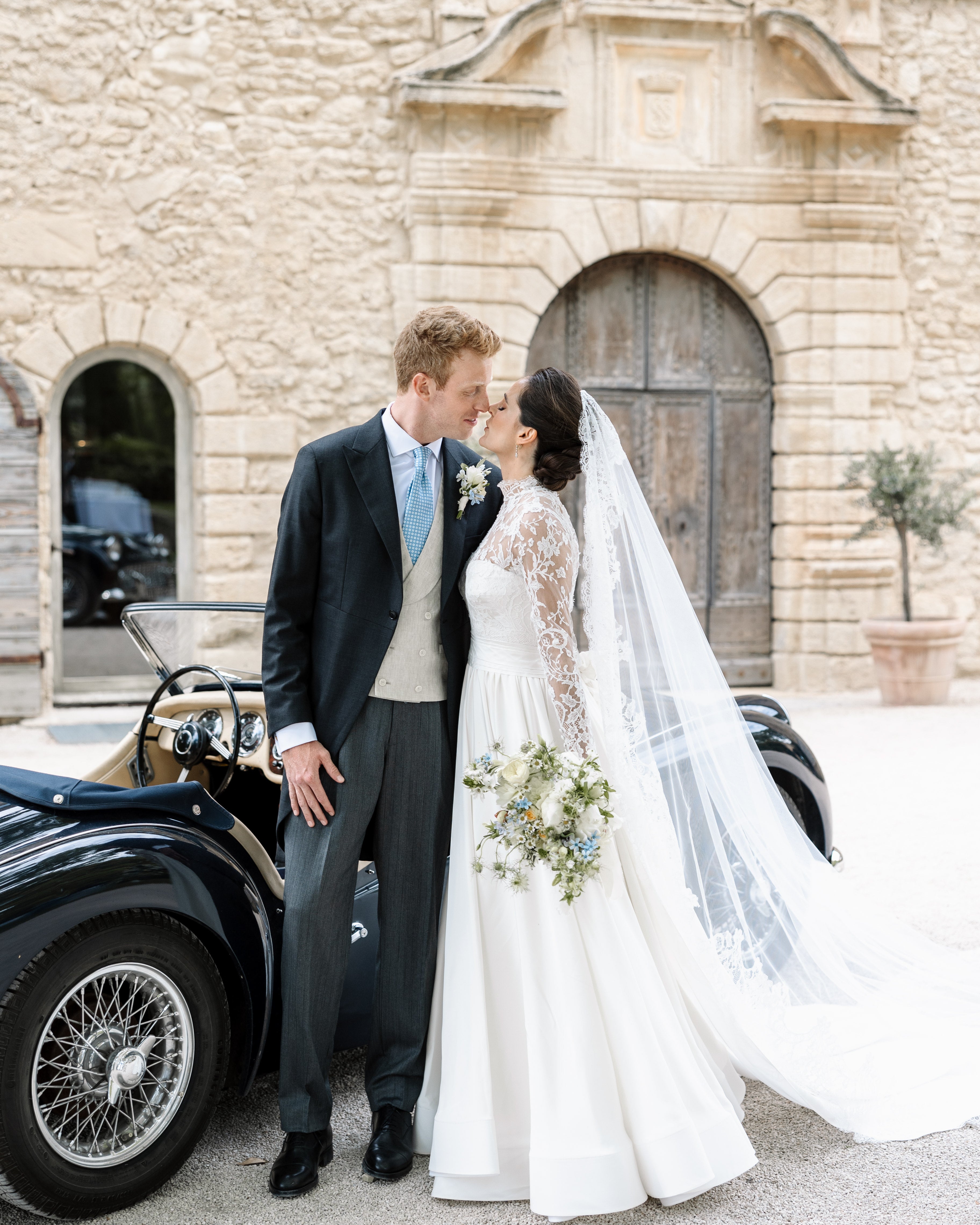 Young bride and groom kissing in front of a French castle and old car after getting married. The bride is  wearing an ivory cathedral bridal veil in fine tulle with elegant lace edge, as a mantilla, and lace wedding dress.